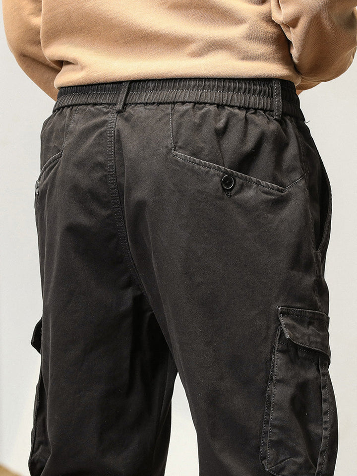 Un homme porte un pantalon cargo noir à poches boutonnées. Coupe droite avec bas resserré aux chevilles. Tailles US 28 à 38. Matières : 97% coton + 3% Spandex. Confortable et flexible. [Cargo District]