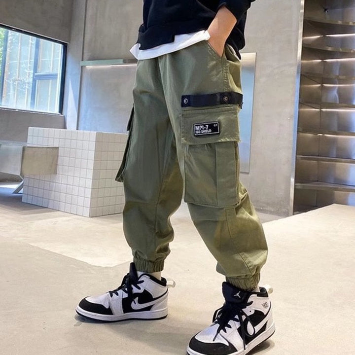 Un garçon porte un pantalon cargo jogger large kaki avec des poches latérales pratiques et des chevilles resserrées. Tailles disponibles du 110 au 160 cm. Style urbain et confort maximal.