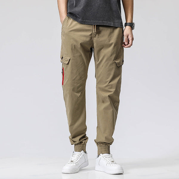 Un homme pose sur fond blanc dans un tee shirt noir, baskets blanches avec une montre et un pantalon cargo coupe droite resserré aux chevilles avec fermeture éclair rouge. Le pantalon est vert olive.