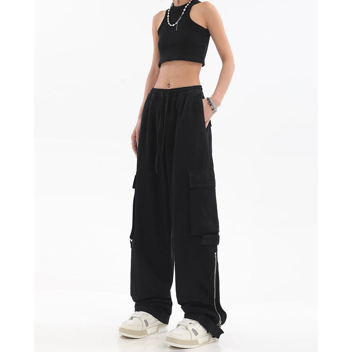 Une femme porte un crop top noir, des baskets blanches ainsi qu'un pantalon cargo baggy noir avec des fermetures éclair sur le bas du pantalon. Elle porte aussi de gros bijoux en argent.