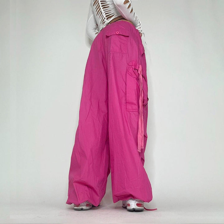 Une femme portant un pantalon cargo baggy rose avec une chemise blanche et des lanières pendantes. Le pantalon offre un confort sans égal avec sa coupe large façon parachute et sa taille basse élastique. Disponible du S au L.