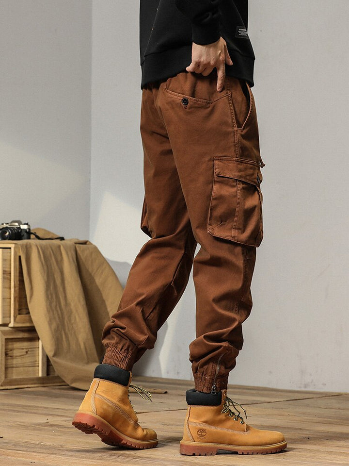 Un homme portant un pantalon cargo camel avec des poches à bouton. Coupe droite et bas resserré aux chevilles. Tailles disponibles du 28 au 38.