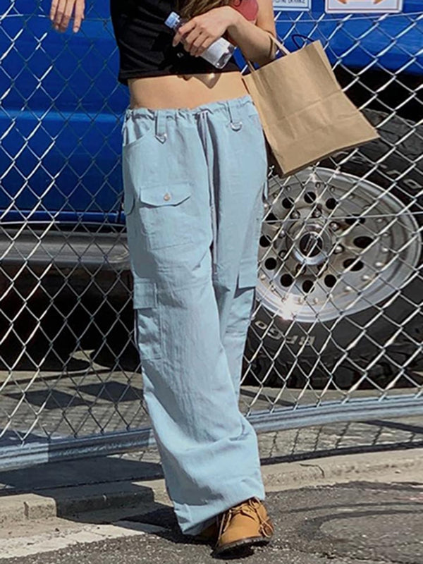 Une femme pose devant un grillage. Elle porte un top court noir, des boots camel, un pantalon cargo taille basse élastique bleu ciel. Elle porte une bouteille d'eau et un sac en papier dans la main.