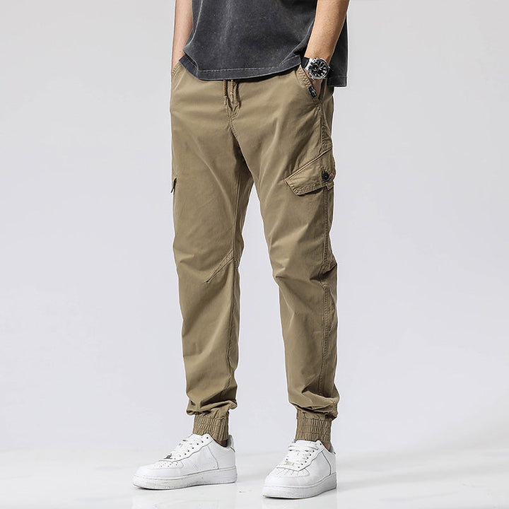 Un homme porte un pantalon cargo coupe droite avec une fermeture rouge, de couleur vert olive. Le pantalon est composé de 95% coton et 5% Spandex, offrant douceur, résistance et souplesse. Disponible du 28 au 38 (tailles US), il présente une coupe droite et resserrée à la cheville pour structurer la silhouette. Une poche est ornée d'une fermeture éclair rouge, ajoutant une touche de couleur audacieuse. Le pantalon est vendu sur Cargo District, un site de vente de vêtements de style cargo.