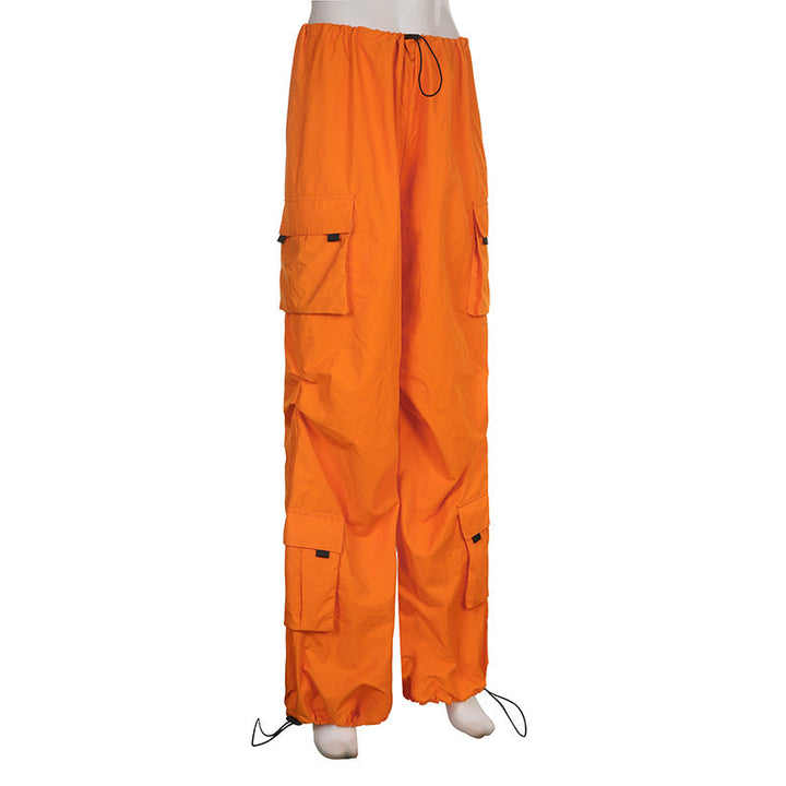 Une personne porte un pantalon orange baggy taille basse pour femme avec des lanières pendantes. Coupe large façon parachute, confortable et extensible. Disponible du S au L. Cargo District, site de vente de vêtements de style cargo.