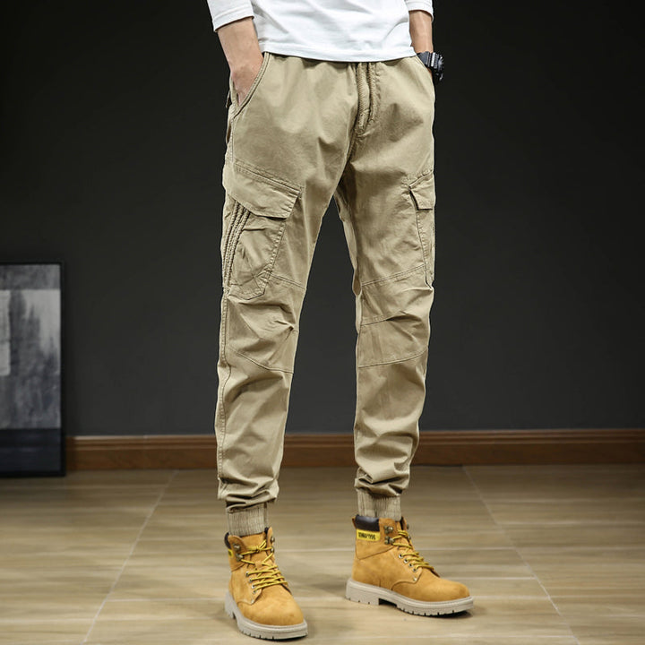 Un homme porte un pantalon cargo slim resserré aux chevilles de couleur beige. Le pantalon est confortable et élégant, avec une coupe slim et des coutures aux genoux. Disponible en tailles US 28 à 38.