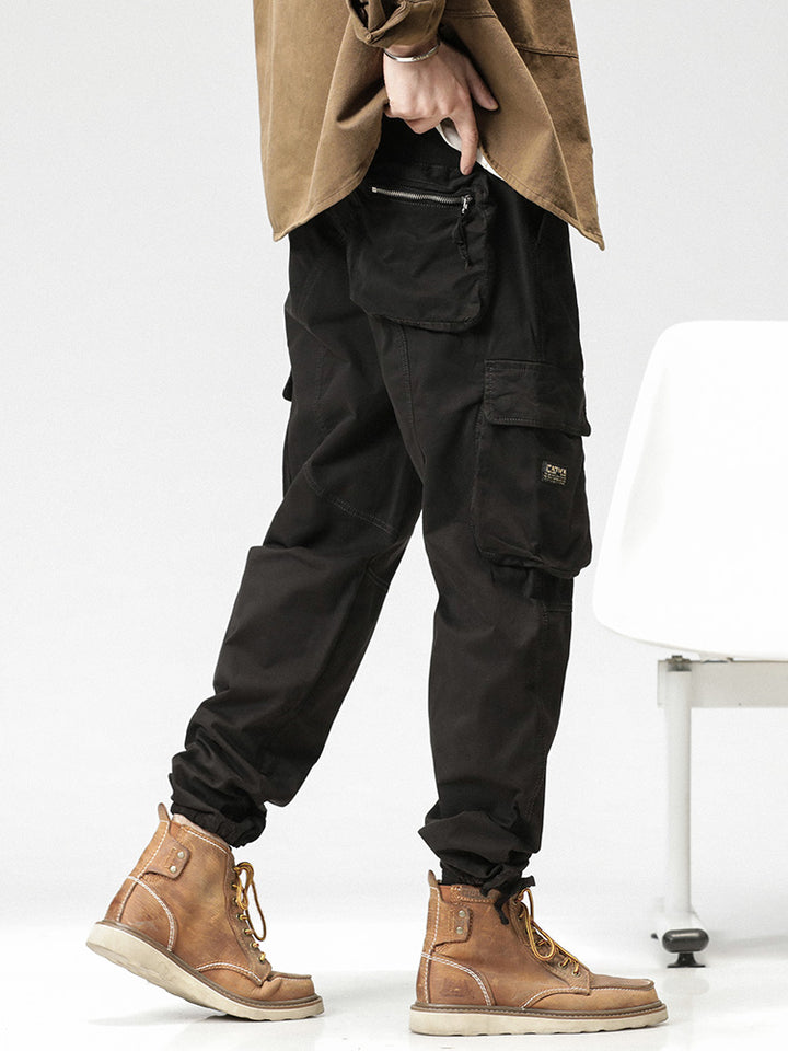 Un homme portant un pantalon cargo noir ample avec des poches latérales et une fermeture bouton et éclair. Le pantalon offre confort et flexibilité grâce à sa composition en coton et spandex. Parfait pour un look décontracté et pratique.