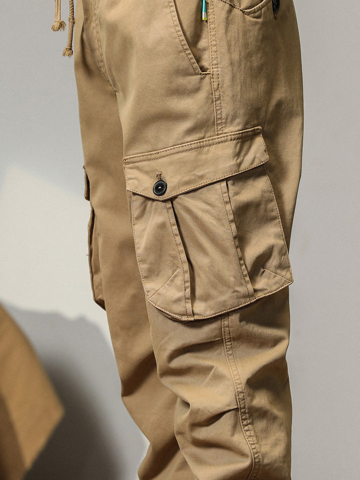 Un pantalon cargo beige pour homme avec poches à bouton, coupe droite. Poches à rabats équipées d'un bouton apparent pour fermeture sécurisée. Bas resserré aux chevilles. Tailles US 28 à 38.