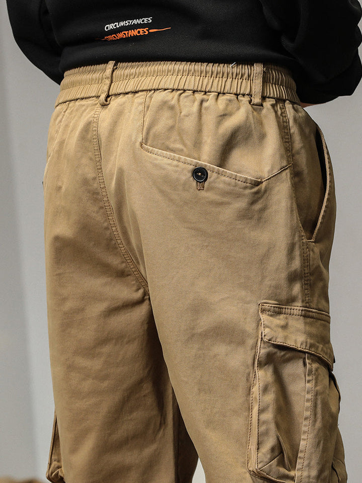 Un homme porte un pantalon cargo beige avec des poches à bouton. Coupe droite et bas resserré aux chevilles. Tailles US 28 à 38.