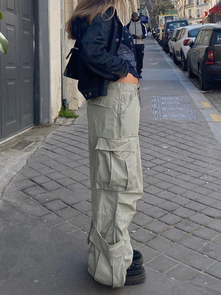 Une femme debout sur un trottoir, portant un pantalon cargo large taille basse gris. Le pantalon a un style baggy avec des poches latérales spacieuses. Disponible du S au L. (Cargo District)