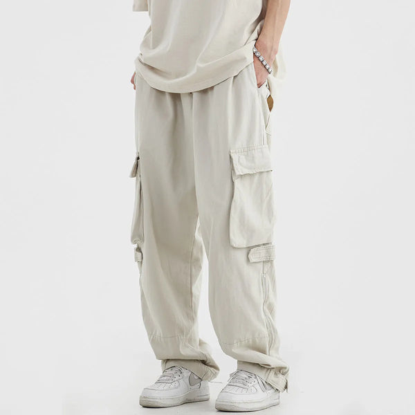 Un homme pose dans un pantalon cargo large beige avec des fermetures éclair sur le côté des chevilles. Il a aussi un tee shirt beige et et des baskets. 