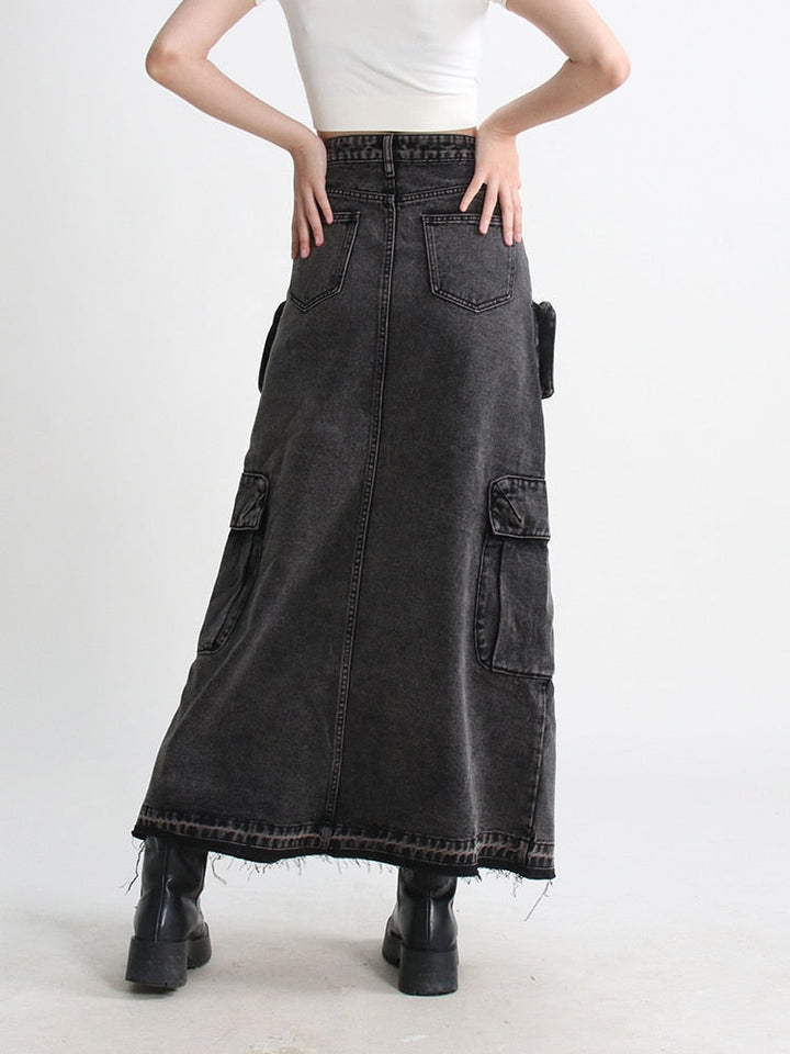 Une femme portant une jupe jean longue fendue multipoches noire, ajoutant une touche urbaine et tendance à son style. La jupe cargo taille haute offre confort et liberté de mouvement avec son design évasé et sa fente. Les nombreuses poches ajoutent une fonctionnalité pratique. Disponible en tailles S à XL.