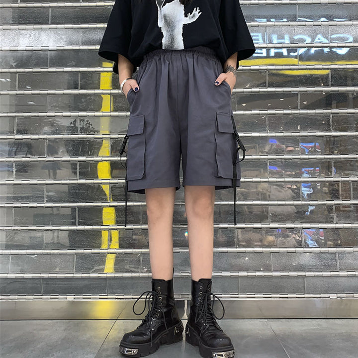 Une femme debout devant un mur, portant un short cargo ample gris avec des poches à clip. Le short est léger, taille haute et arrive au-dessus du genou. Parfait pour un style urbain décontracté. Disponible en tailles du S au 4XL.