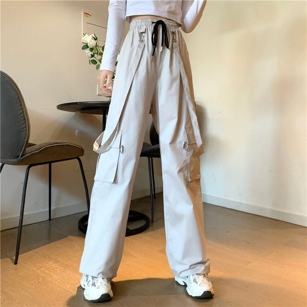 Une femme pose dans une salle à manger avec une table et des chaises derrière elle. Elle porte un tee-shirt manches longues blanc, des baskets blanches ainsi qu'un pantalon cargo taille haute beige avec des sangles ajustables qui peuvent se mettre en bretelles.