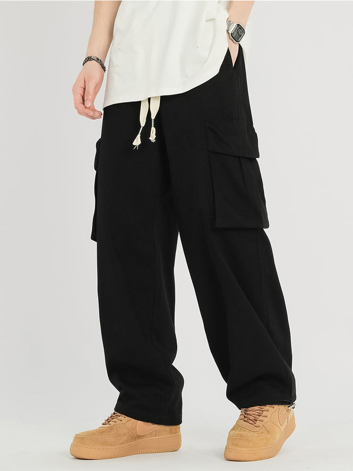 Un homme pose en pantalon cargo baggy en coton noir. Il porte également des baskets camel et un tee-shirt blanc. 
