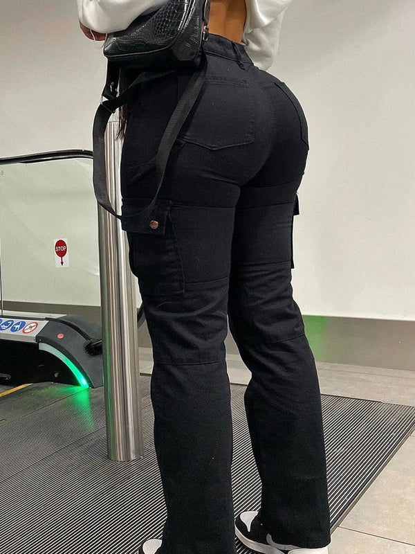 Une femme pose à côté d'un escalator. Elle porte un crop top à manches longues blanc, un sac noir, des baskets noires et blanches ainsi qu'un pantalon cargo noir, taille haute. La coupe du pantalon est ajusté. Elle pose de dos.