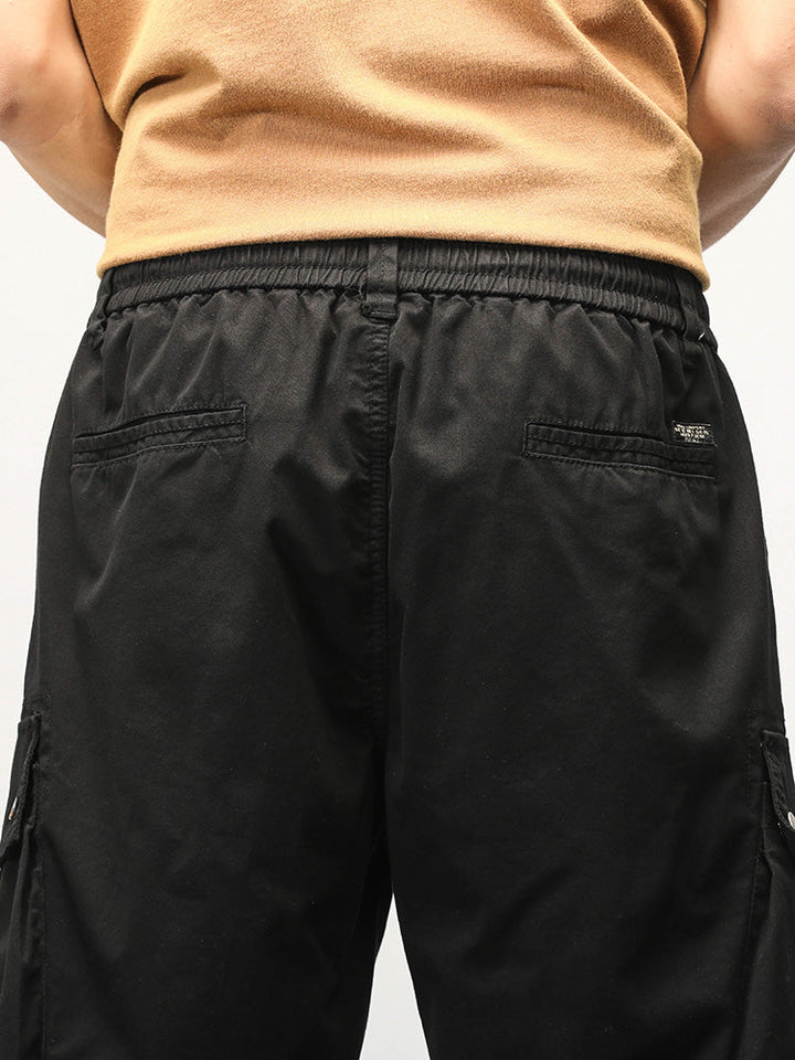 Un homme porte un Short cargo coupe droite noir avec des poches latérales à boutons-pression. Fabriqué à partir de 95% coton et 5% spandex, ce short offre confort et élasticité. Tailles disponibles du 28 au 40.