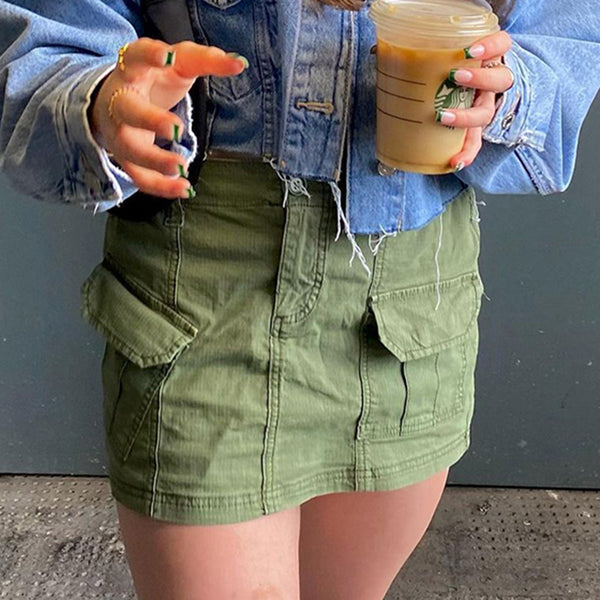 Une femme pose dans la rue avec un café à la main. Elle porte une veste en jean courte ainsi qu'une mini jupe cargo légère kaki. Les poches sont asymétriques.