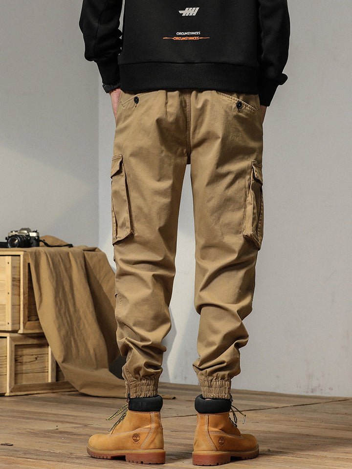 Un homme porte un pantalon cargo beige avec des poches à bouton. Coupe droite et bas resserré aux chevilles. Élastique à la taille et cordon pour ajustement parfait. Disponible en tailles US 28 à 38.