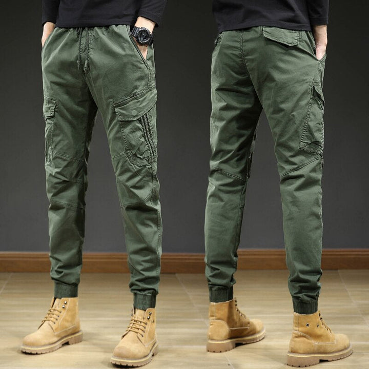 Un homme portant un pantalon cargo slim kaki avec des chevilles resserrées. Confortable et élégant, ce pantalon est doté d'une taille élastique avec cordon pour un ajustement parfait. Sa coupe slim et ses coutures aux genoux créent un look moderne. Disponible en tailles US 28 à 38.