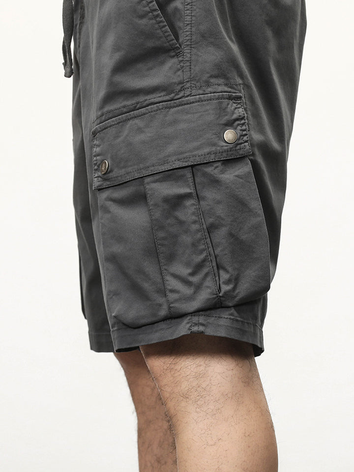 Un homme portant un short cargo coupe droite gris avec des poches latérales à boutons-pression. Fabriqué à partir de 95% coton et 5% spandex, ce short offre un confort maximal et une légère élasticité. Tailles disponibles du 28 au 40.