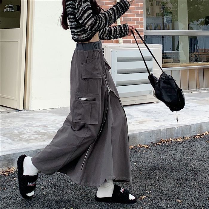 Une femme marchant dans la rue avec une jupe cargo longue zippée grise et un sac à dos noir.