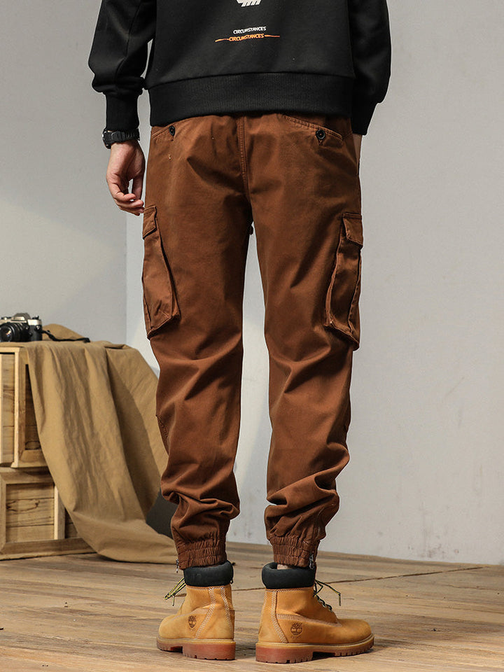 Un homme porte un pantalon cargo coupe droite de couleur camel avec des poches à bouton. Le bas du pantalon est resserré aux chevilles pour un look masculin. Disponible en tailles US 28 à 38.