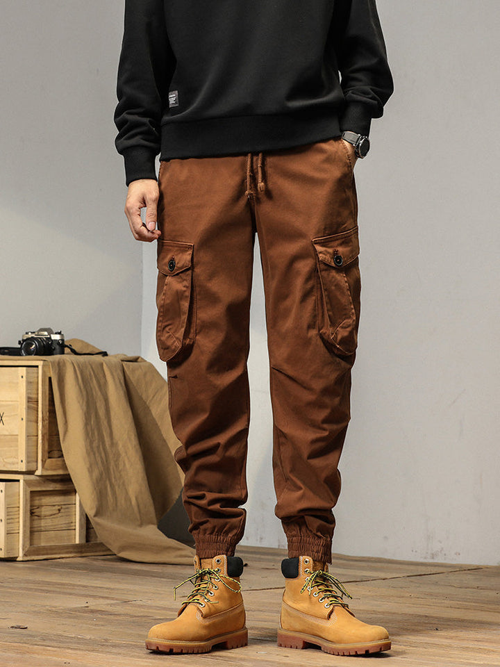 Un homme portant un pantalon cargo camel avec poches à bouton. Coupe droite et bas resserré aux chevilles. Élastique à la taille et cordon pour un ajustement parfait. Disponible en tailles US 28 à 38.