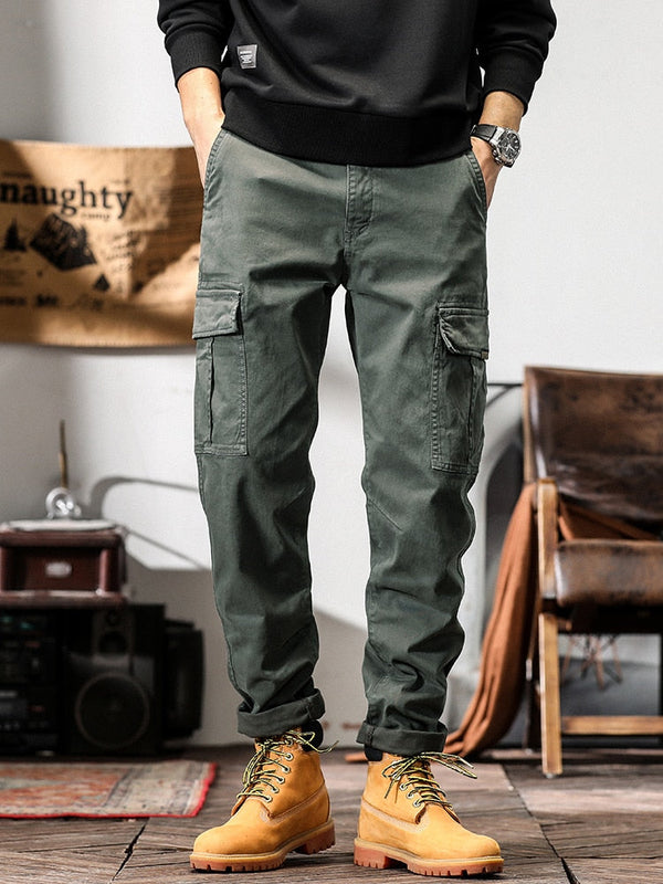 Un homme pose dans un décor industriel avec un sweat noir, une montre et des boots marrons. Il porte également un pantalon cargo coupe droite gris.