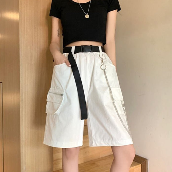 Une femme pose dans des escaliers. Elle porte un tee-shirt noir court ainsi qu'un short long cargo blanc avec ceinture noire et chaîne.