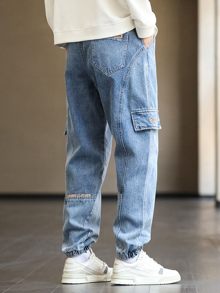 Un homme portant un jean cargo jogger bleu avec élastique à la taille et cordon bicolore blanc et turquoise. Coupe légèrement large avec patchs marron et coutures structurantes. Disponible du M au 8XL.