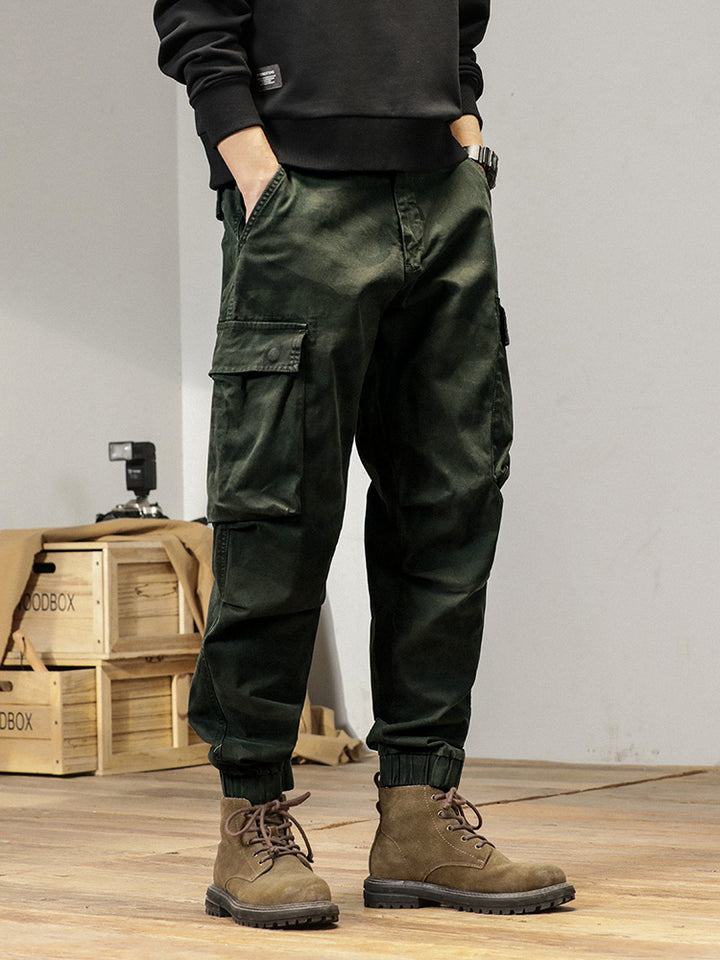 Un homme porte un pantalon cargo kaki avec une coupe large resserrée aux chevilles. Le pantalon est fait de 100% coton pour un confort optimal et une durabilité exceptionnelle. Disponible en tailles US 28 à 38.