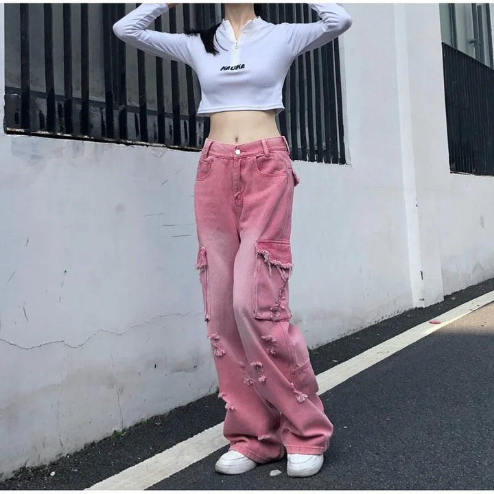 Une femme debout devant un bâtiment blanc, portant un jean cargo baggy rose délavé avec des déchirures. Taille haute pour une silhouette moderne. Un look audacieux des années 2000. Disponible en tailles XS à XL.