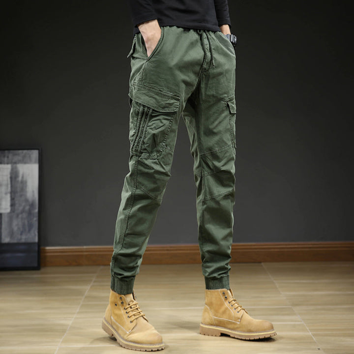 Un homme porte un pantalon cargo slim resserré aux chevilles de couleur kaki. Les coutures aux genoux et au bas resserré créent un look moderne. Le pantalon est confortable et flexible grâce à sa composition en coton (95%) et élasthanne (5%). Disponible en tailles US 28 à 38.