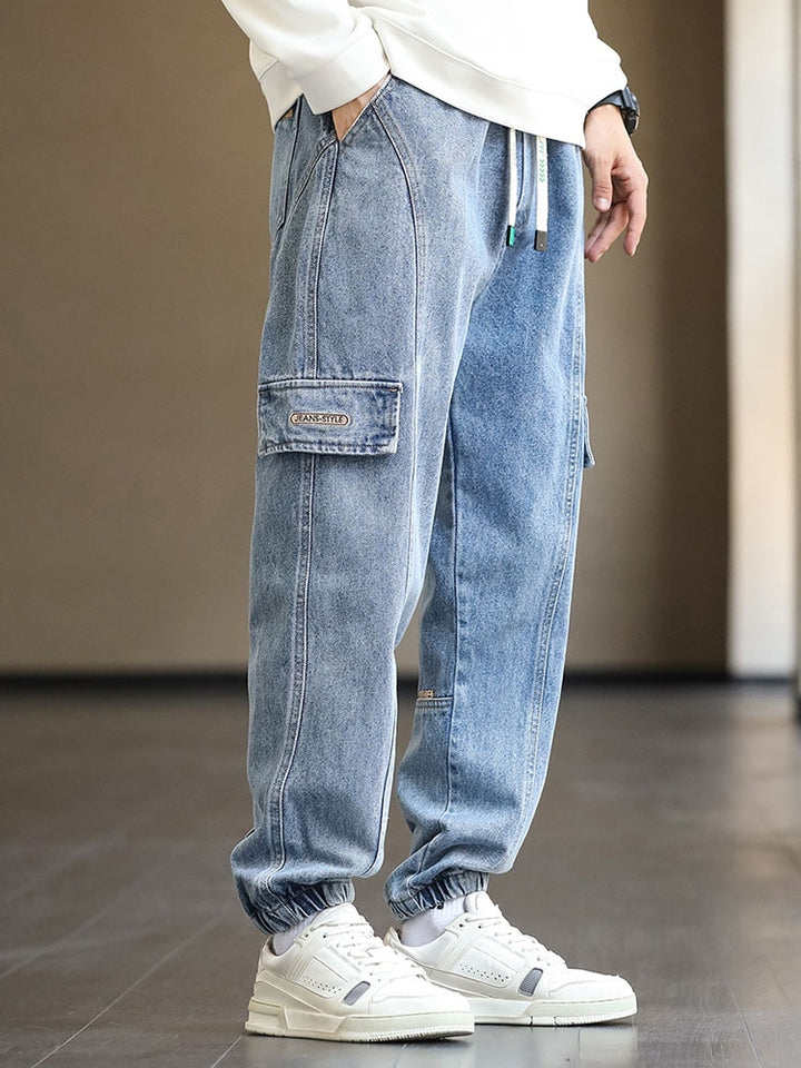 Un homme portant un jean cargo jogger bleu avec des sneakers blancs. Le jean a une taille élastique et un cordon bicolore blanc et turquoise pour un ajustement optimal et une touche de style. Coupe légèrement large avec des patchs marron et des coutures structurantes pour une allure street tendance. Disponible du M au 8XL.