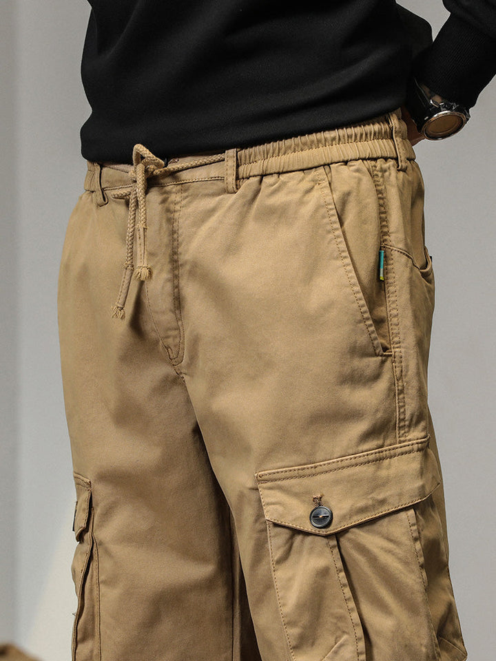 Un homme porte un pantalon cargo beige avec des poches à bouton. Coupe droite et bas resserré aux chevilles, il offre un look masculin. Disponible en tailles US 28 à 38.