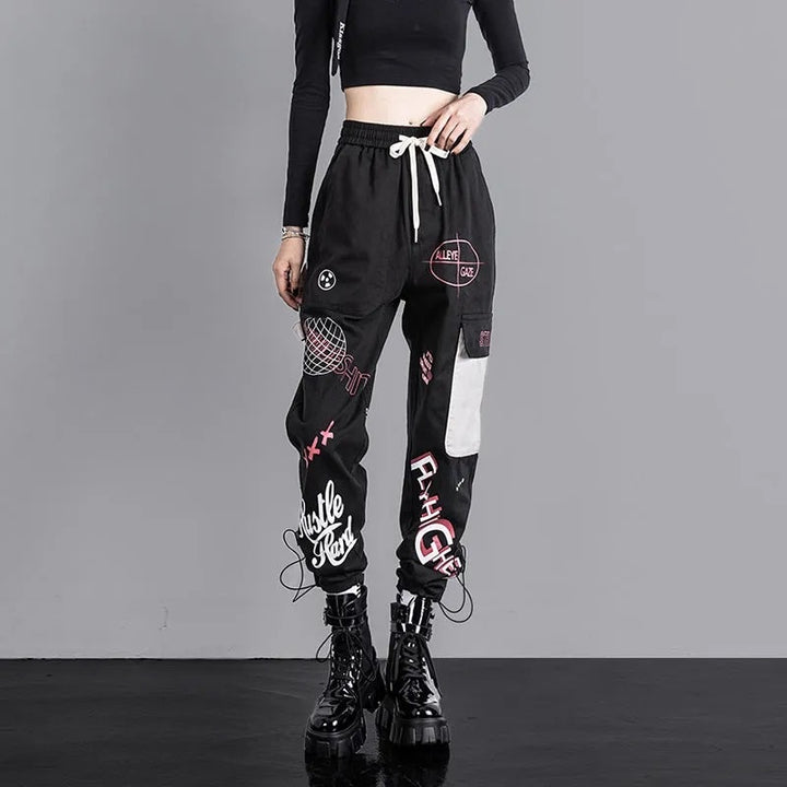 Une femme porte un Pantalon cargo jogger noir à motif avec des patches. Ce pantalon cargo noir a une coupe jogger taille haute, des motifs blancs et roses, une poche latérale et une lanière gris clair pour une allure urbaine. Disponible du S au 3XL, en coton pour un confort optimal.