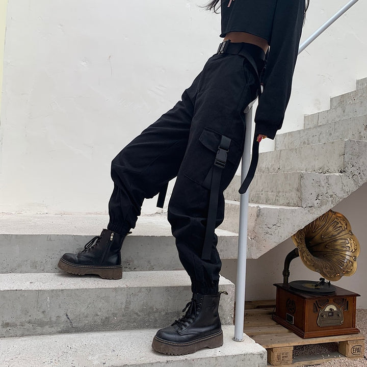 Une femme pose dans des escaliers en béton avec un pull court noir, des boots plateformes noirs et un cargo taille haute resserré aux cheville avec des poches qui se ferment par des clips en plastique. Le pantalon est noir. Il y a un vieux tourne-disque sous l'escalier.