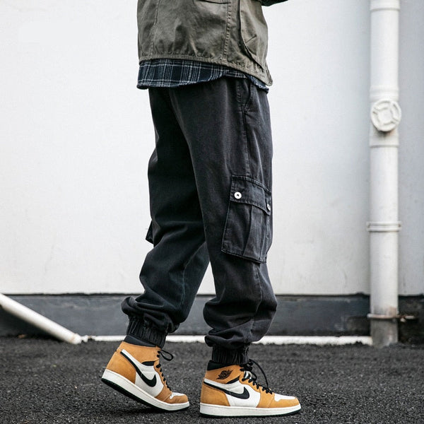 Un homme pose dans la rue avec des baskets camel, blanches et noires. Il porte aussi une chemise à carreaux, une veste kaki et un pantalon jogger coupe large noir.