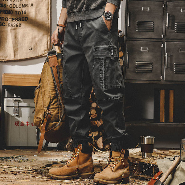 Un homme pose dans un décor industriel avec un pantalon cargo ample resserré aux chevilles noir. Il porte des boots, style Timberland, aux pieds. Il porte un sweat gris foncé, une montre noire et un gros sac à dos de randonneur dans la main.