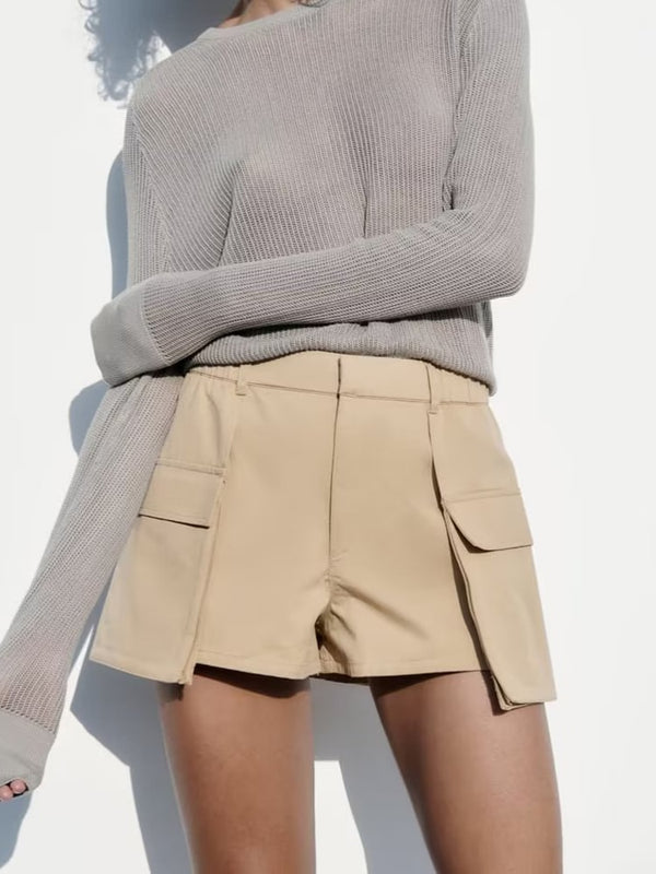 Une femme à la peau halée pose derrière un mur blanc. Elle porte un pull gris et une mini jupe short cargo beige. Les poches latérales sont plus longues que la jupe elle-même.