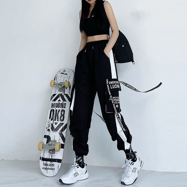 Une femme pose dans une pièce blanche. Elle pose à côté d'un skateboard. Elle porte un top court noir, un pantalon cargo jogger noir avec lanières à inscriptions et bandes latérales blanches. Elle porte aussi des baskets blanches. 