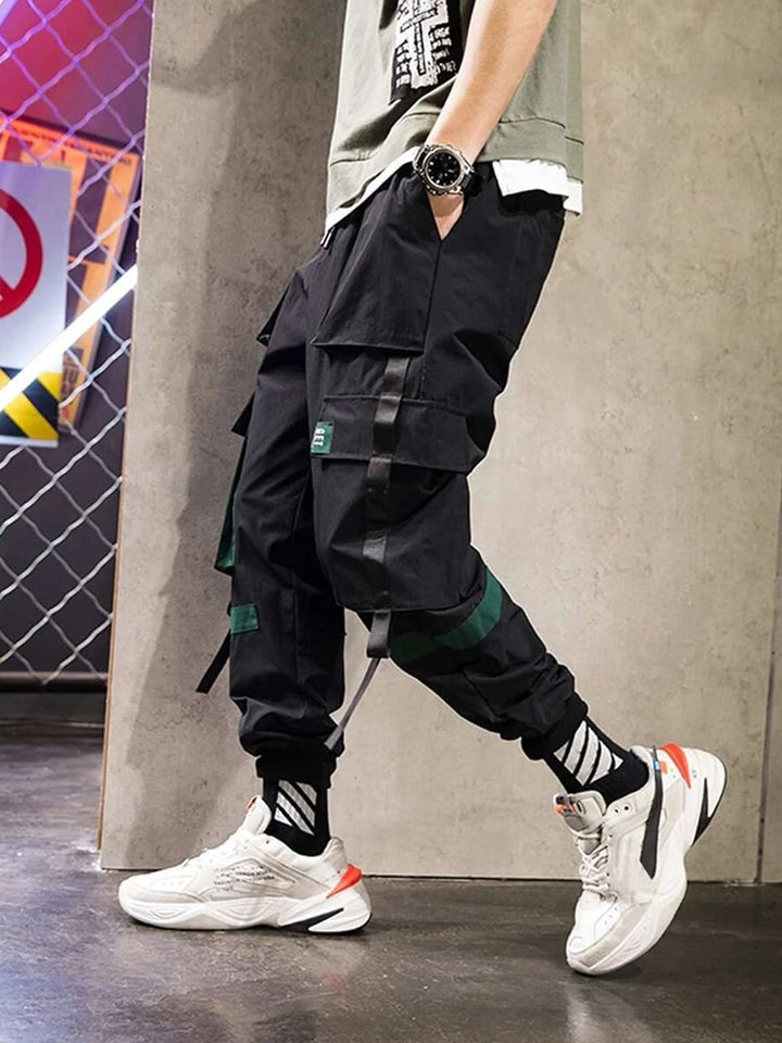 Un homme pose dans un parking avec un tee-shirt kaki, des baskets blanches et un pantalon cargo à lanières ample noir. 