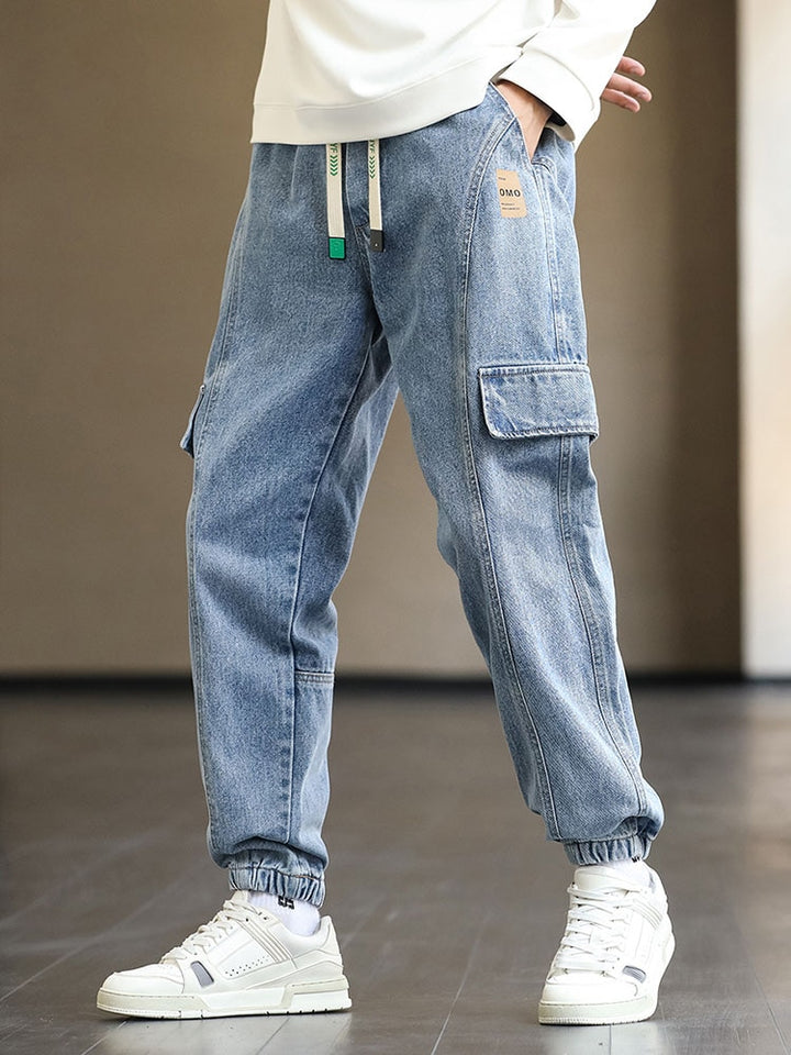 Un homme pose dans une pièce au sol gris et murs beiges. Il porte un sweat blanc, des baskets blanches ainsi qu'un jean jogger bleu avec cordon blanc et turquoise.