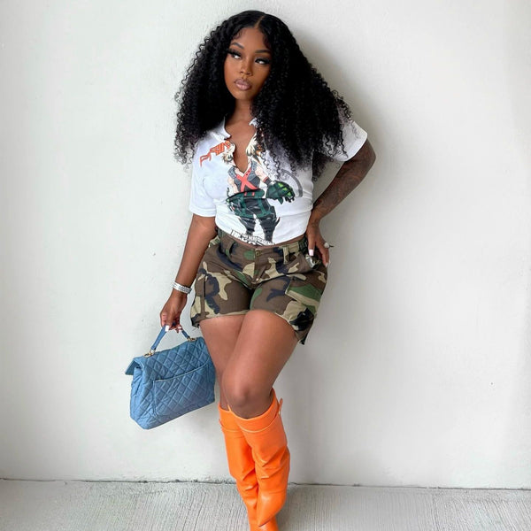 Une femme pose devant un mur blanc. Elle porte un tee-shirt blanc avec un motif, un sac en jean matelassé, des bottes oranges et un short cargo ajusté taille haute vert camo.