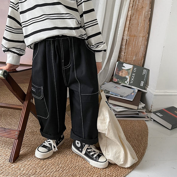 Un enfant pose dans un salon. Il y a un tapis rond en jute, des magazines, un tabouret. L'enfant porte une marinière blanche à rayures noires, des baskets montantes noires et blanches. Il porte un pantalon cargo noir ample avec des coutures contrastantes blanches.