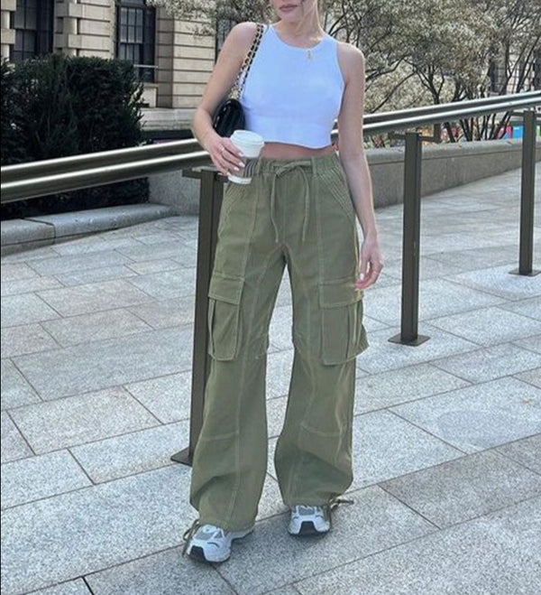 Une femme pose dans la rue avec un café à la main. Elle porte un sac à main noir, un top court blanc, des baskets et un pantalon cargo vert à coutures blanches. Le pantalon est large.