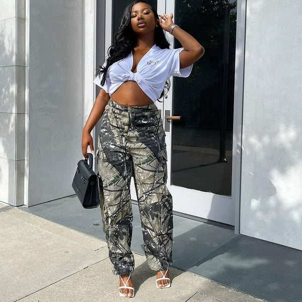 Une femme aux cheveux longs pose dans la rue avec un tee-shirt blanc, des sandales à talons blanches ainsi qu'un sac noir et un pantalon cargo taille haute ajustée vert avec un imprimé feuilles. 